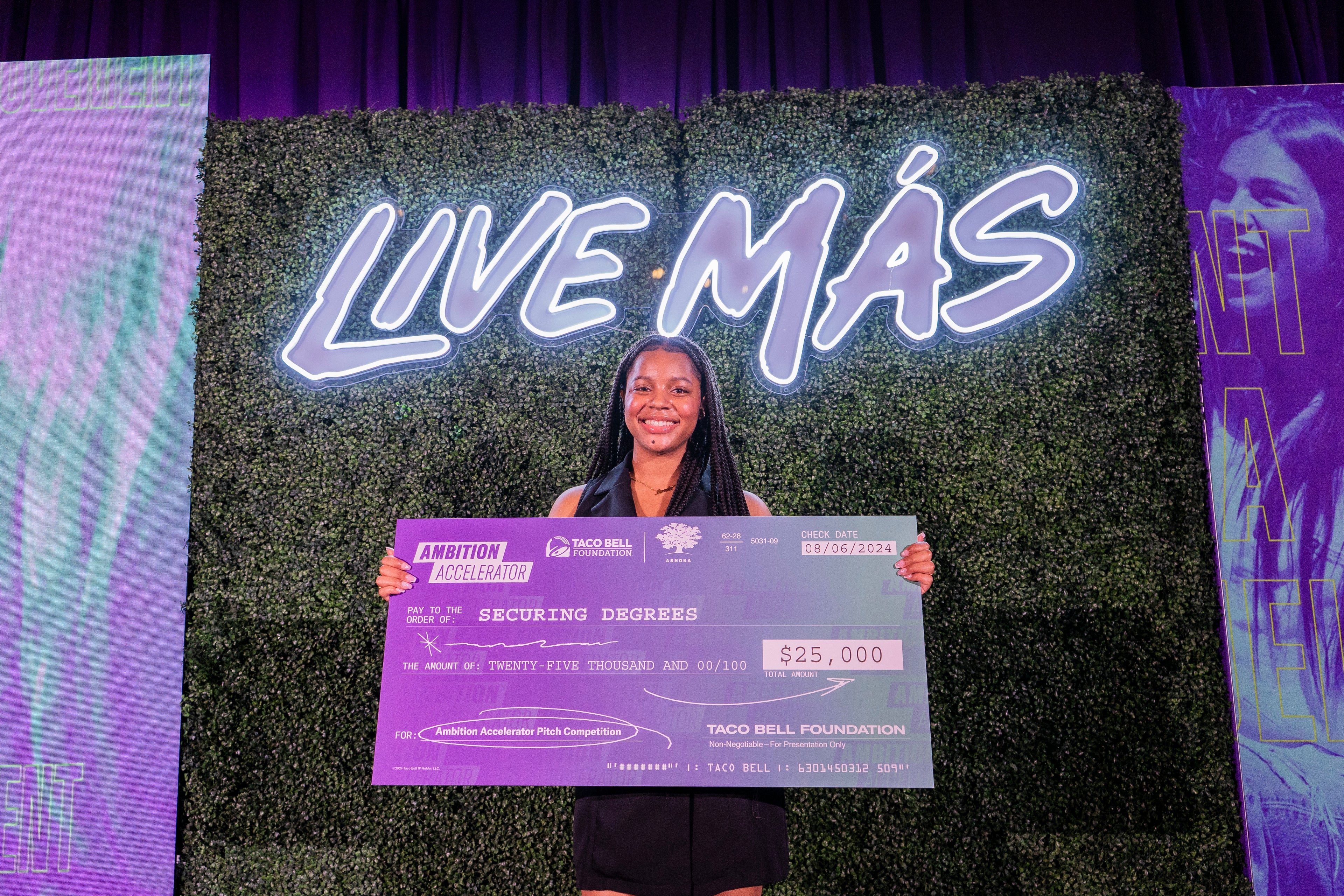 A smiling woman stands in front of a “LIVE MÁS” sign holding an oversized check for $25,000 from the Taco Bell Foundation, awarded for the Securing Degrees project at the Ambition Accelerator event.