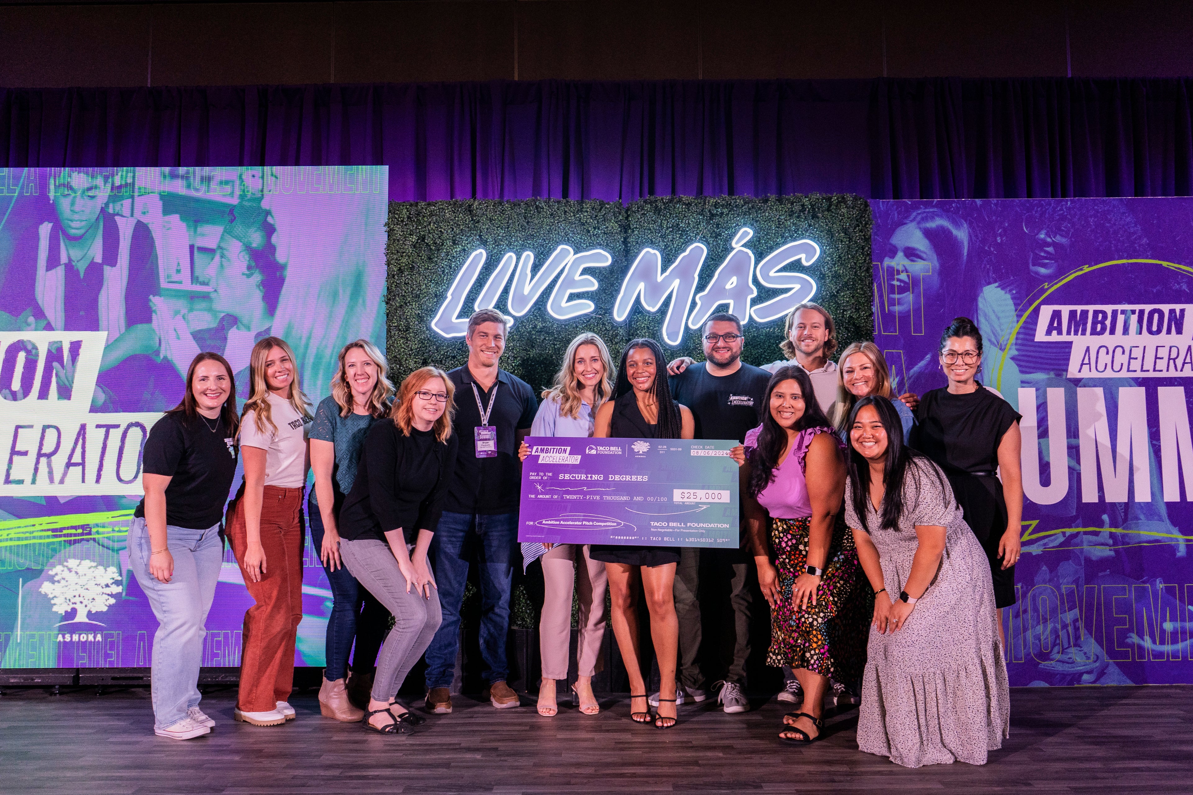 A diverse group of people stand smiling on a stage holding an oversized check. Behind them, a neon sign reads LIVE MÁS and colorful banners promote an accelerator event.