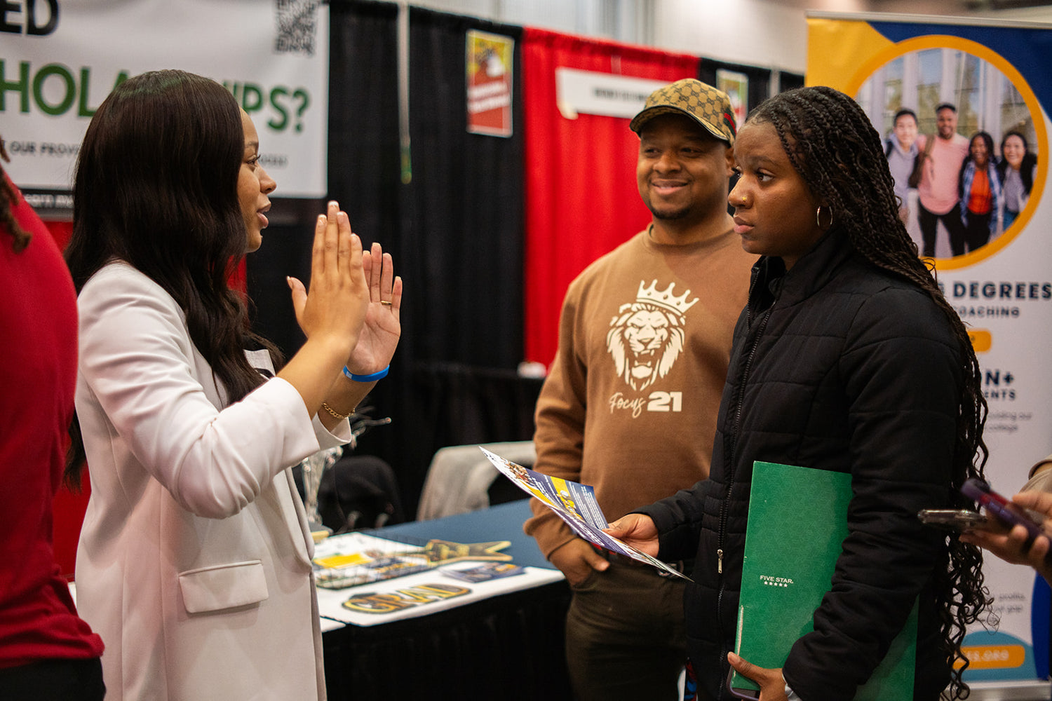 A woman in a white jacket gestures while speaking to two young adults at a college fair booth. The young woman holds brochures and a green folder; the young man wears a tan cap and brown sweatshirt with a lion graphic.