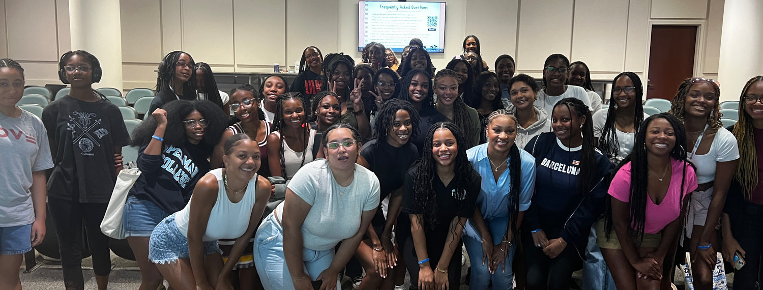 A large group of smiling young women poses together in a classroom or lecture hall, with chairs and a presentation screen visible in the background.
