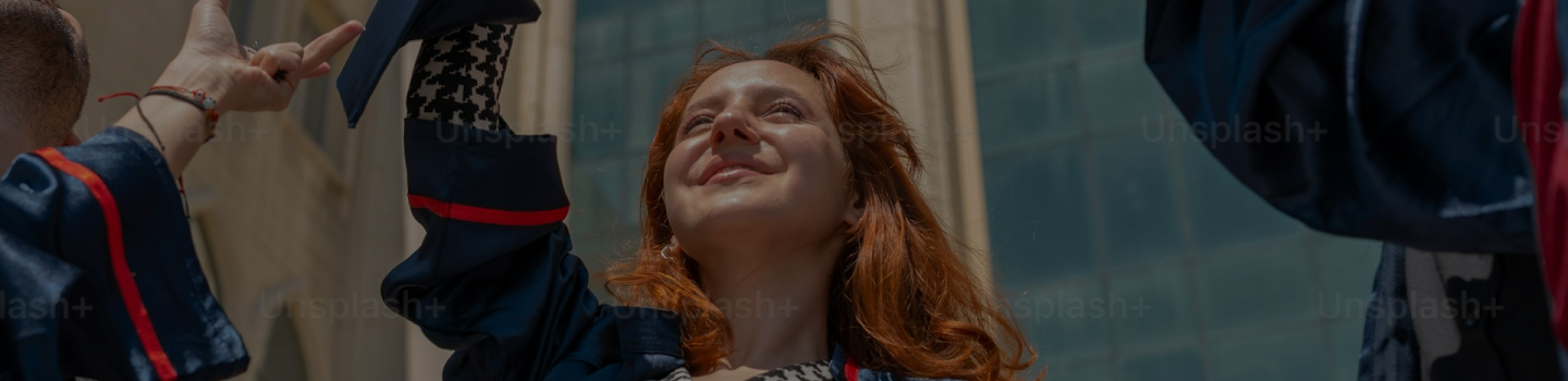 A young woman in graduation attire smiles and looks upward, celebrating outdoors in sunlight with a modern building in the background. Other graduates are partially visible beside her.