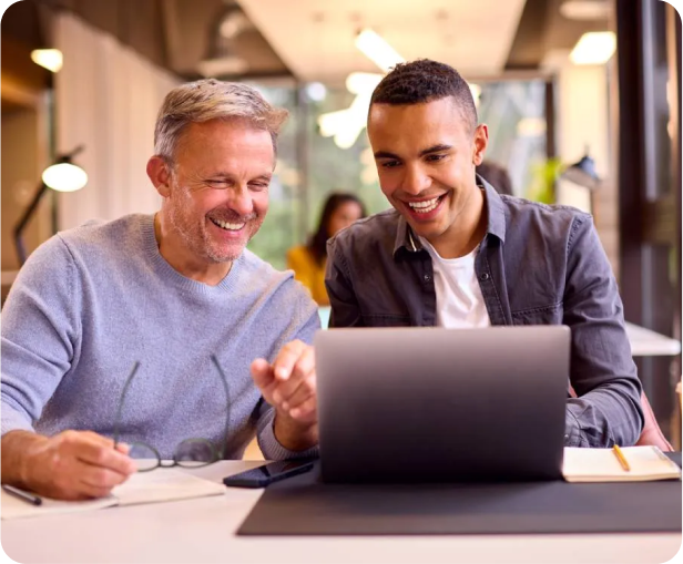 Two men sit together at a table, smiling and looking at a laptop screen. One holds eyeglasses and a notebook is nearby. The setting appears to be a bright, modern office or coworking space.