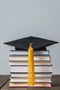 A black graduation cap with a yellow tassel rests on top of a stack of hardcover books, placed on a wooden surface against a plain light gray background.