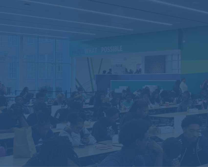 A large group of students sits at long tables in a brightly lit cafeteria, eating, talking, and using their phones. The back wall displays the phrase “PROVE WHAT’S POSSIBLE” in bold letters.