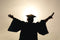 A silhouette of a graduate in cap and gown stands with arms outstretched, holding a diploma, against a bright, sunlit sky.