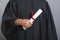 A person wearing a black graduation gown holds a rolled-up diploma tied with a red ribbon against a plain gray background.