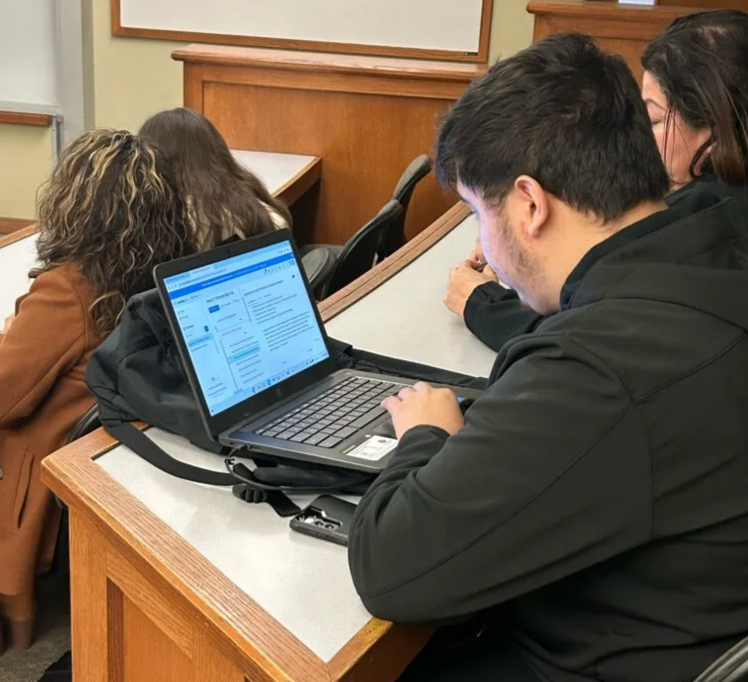 A student sits in a classroom taking notes on a laptop, with other students seated nearby, all facing forward toward the instructor or whiteboard.