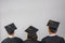 Three graduates wearing black caps and gowns are seen from behind, standing in front of a plain gray background. The yellow tassels on their caps are hanging to the right.