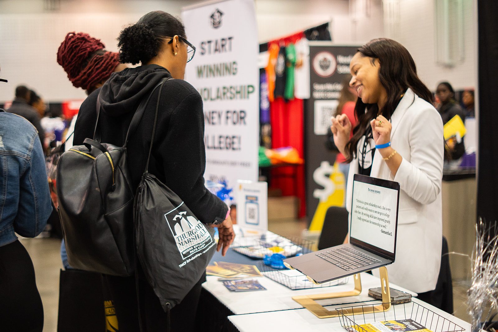 A woman at an information booth smiles and talks to two visitors at an event. A laptop, flyers, and a sign about winning scholarship money for college are on the table. Colorful banners and people are in the background.