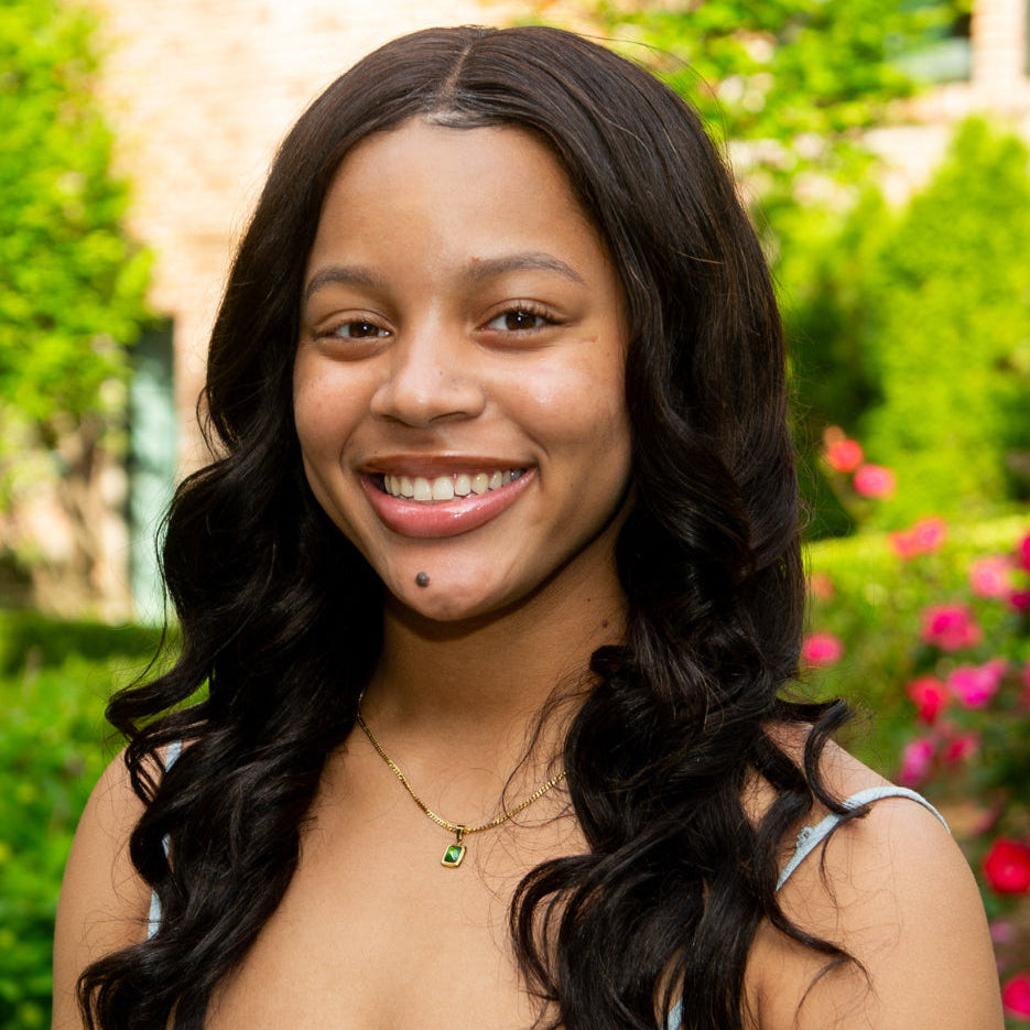 A young woman with long, wavy dark hair smiles outdoors. She wears a gold necklace and a light-colored top. Green trees and pink flowers are blurred in the background.
