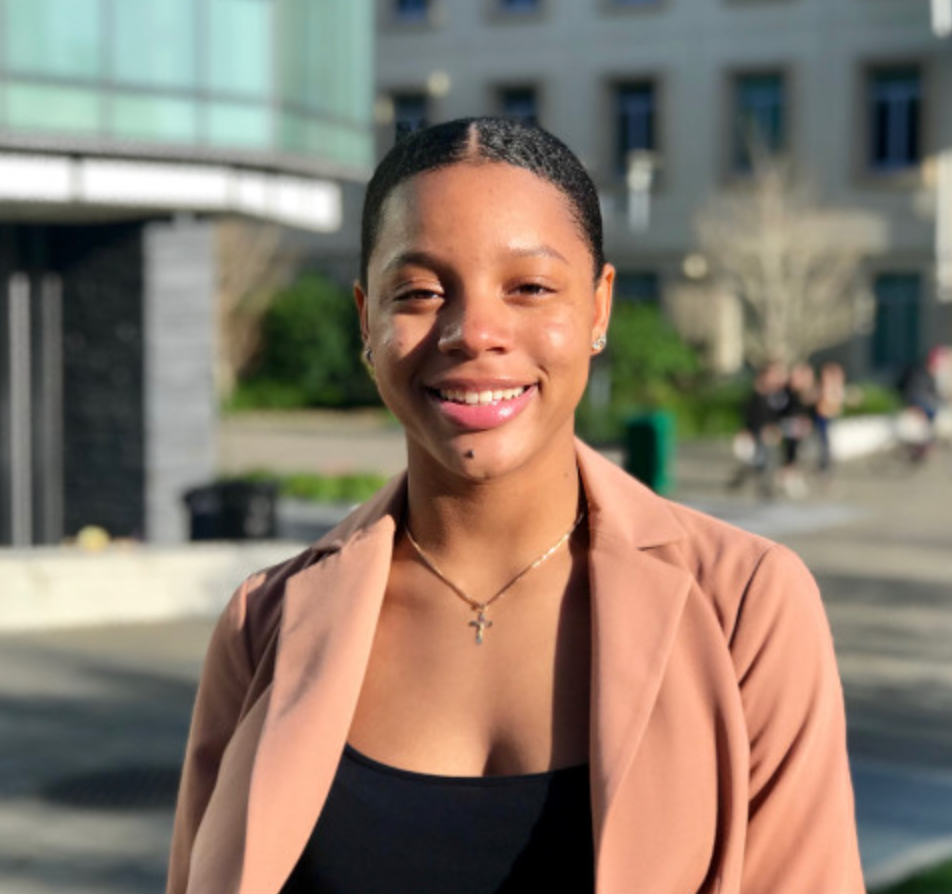 A young woman wearing a light brown blazer and a cross necklace smiles outdoors on a sunny day, with blurred buildings and greenery in the background.