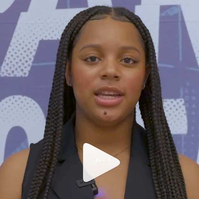 A young woman with long braided hair speaks on camera, wearing a black sleeveless top, standing in front of a purple background with large white letters. A play button icon appears over the image.
