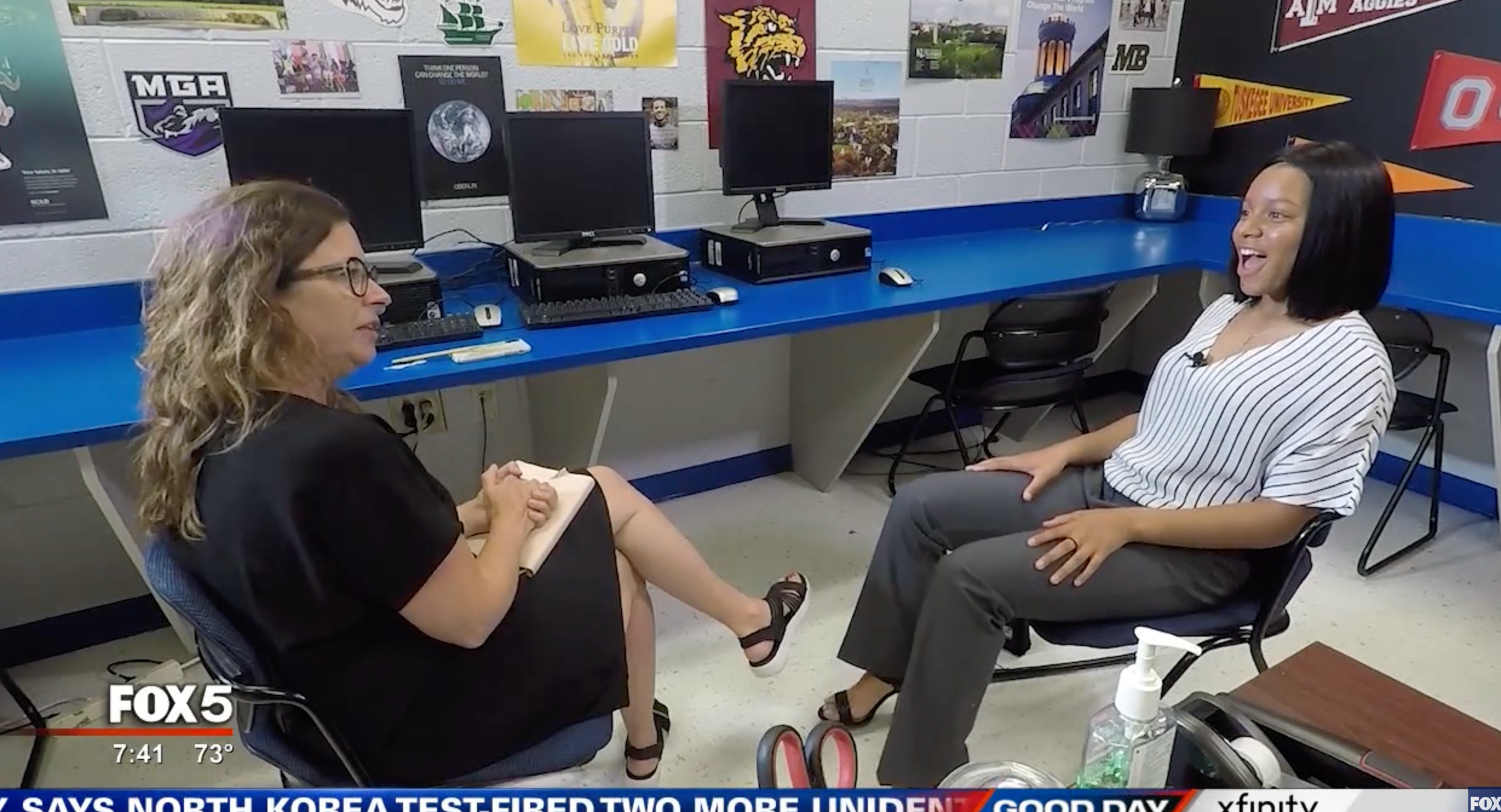 Two women sit across from each other in a computer lab, engaged in conversation. One wears glasses and a black dress, while the other smiles, wearing a white top and gray pants. Various posters decorate the wall behind them.