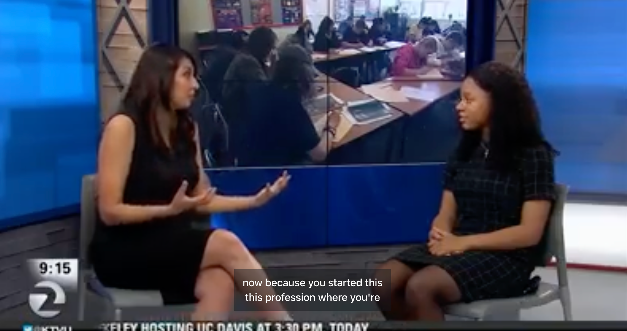 Two women sit and talk in a TV studio with a classroom scene shown on a large screen behind them. One woman gestures with her hands while the other listens. Captions and a news ticker appear at the bottom.